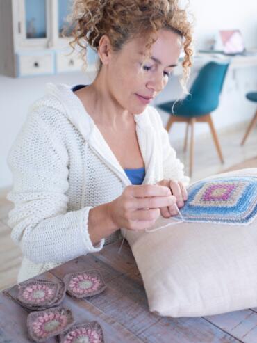 Happy adult woman sewing a pillow with handmade crochet squares sitting comfortably